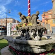 A stone fountain adorned with sculptures of figures, flowers, and a striped pole, set against a blue sky in a lively urban square.