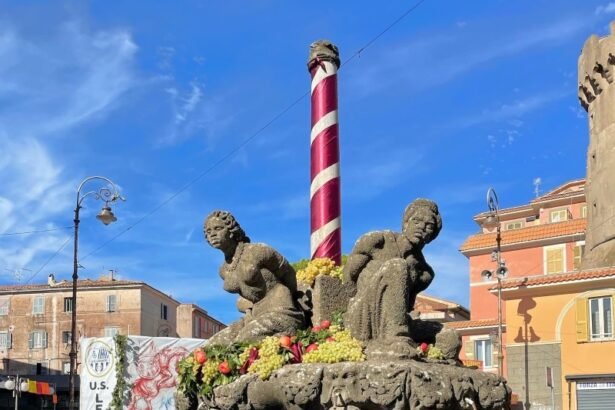 A stone fountain adorned with sculptures of figures, flowers, and a striped pole, set against a blue sky in a lively urban square.