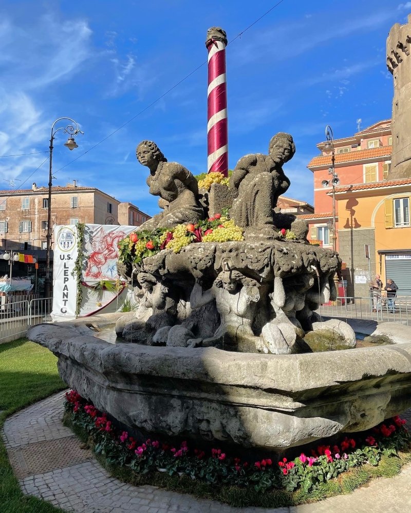 A stone fountain adorned with sculptures of figures, flowers, and a striped pole, set against a blue sky in a lively urban square.