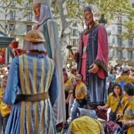 A vibrant festival scene featuring towering giant puppets being paraded among onlookers, dressed in traditional attire, with a crowd of people in colorful outfits enjoying the festivities.