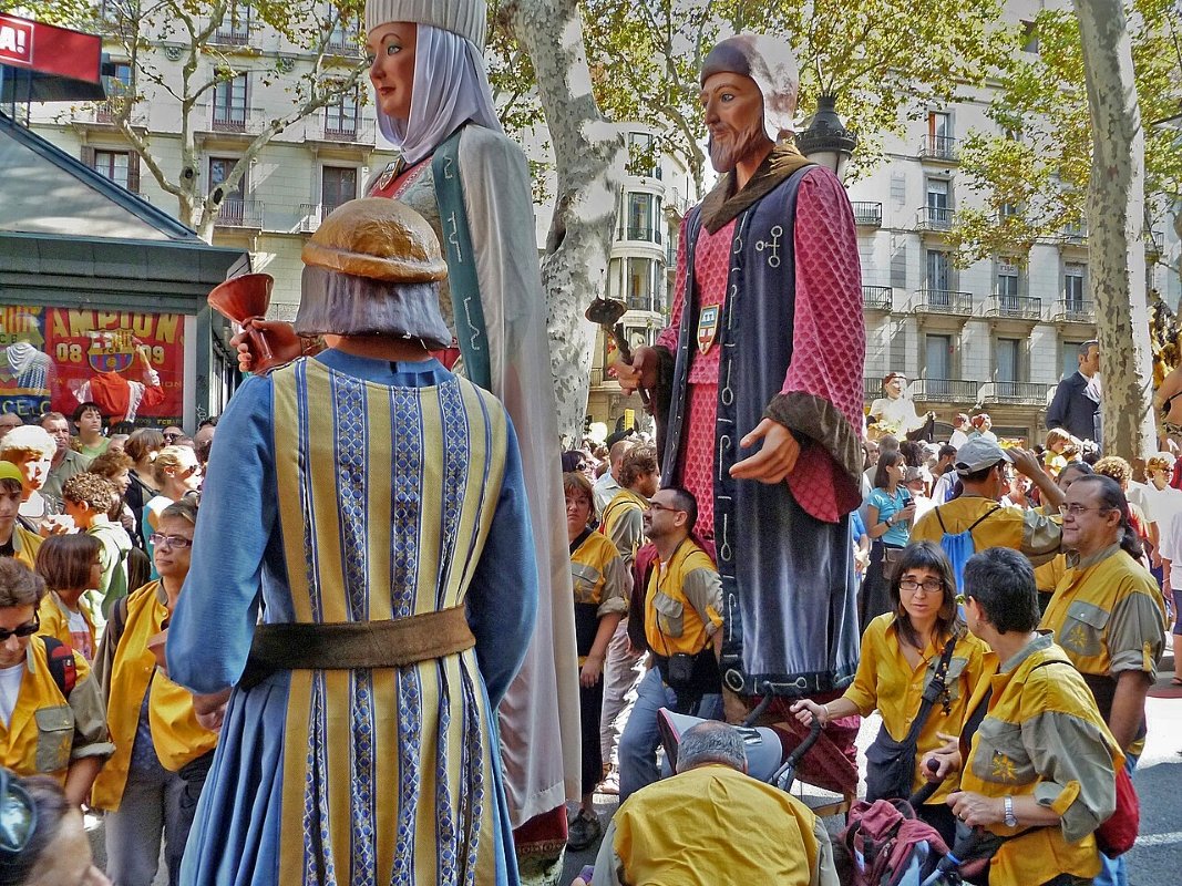 A vibrant festival scene featuring towering giant puppets being paraded among onlookers, dressed in traditional attire, with a crowd of people in colorful outfits enjoying the festivities.