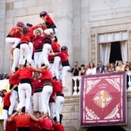 A group of performers constructing a traditional human tower, known as a "castell," in front of the Ajuntament de Barcelona, with spectators watching from a balcony above.
