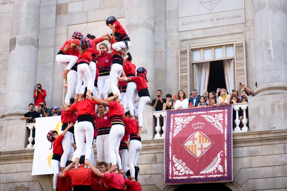 A group of performers constructing a traditional human tower, known as a "castell," in front of the Ajuntament de Barcelona, with spectators watching from a balcony above.