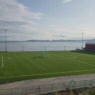 A panoramic view of a well-maintained soccer field adjacent to a tranquil sea, with distant islands visible under a partly cloudy sky.