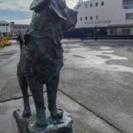 A bronze statue of a dog wearing a hat stands prominently at a harbor, with a ship named "Ocean Explorer" docked in the background and people walking nearby.