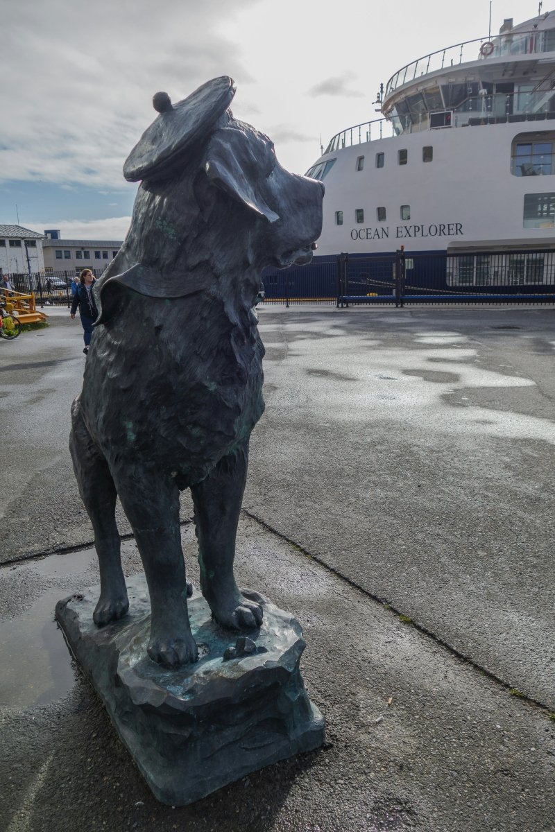 A bronze statue of a dog wearing a hat stands prominently at a harbor, with a ship named "Ocean Explorer" docked in the background and people walking nearby.