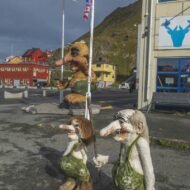 A display of whimsical troll sculptures standing in a parking area, with colorful buildings and flags in the background.