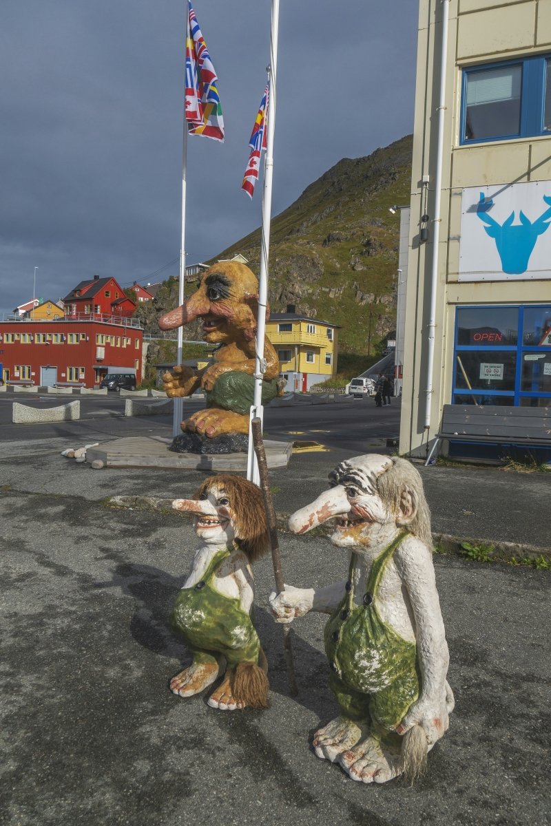 A display of whimsical troll sculptures standing in a parking area, with colorful buildings and flags in the background.