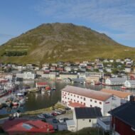 A picturesque view of a coastal town with colorful buildings, boats in the harbor, and a mountain in the background under a clear blue sky.