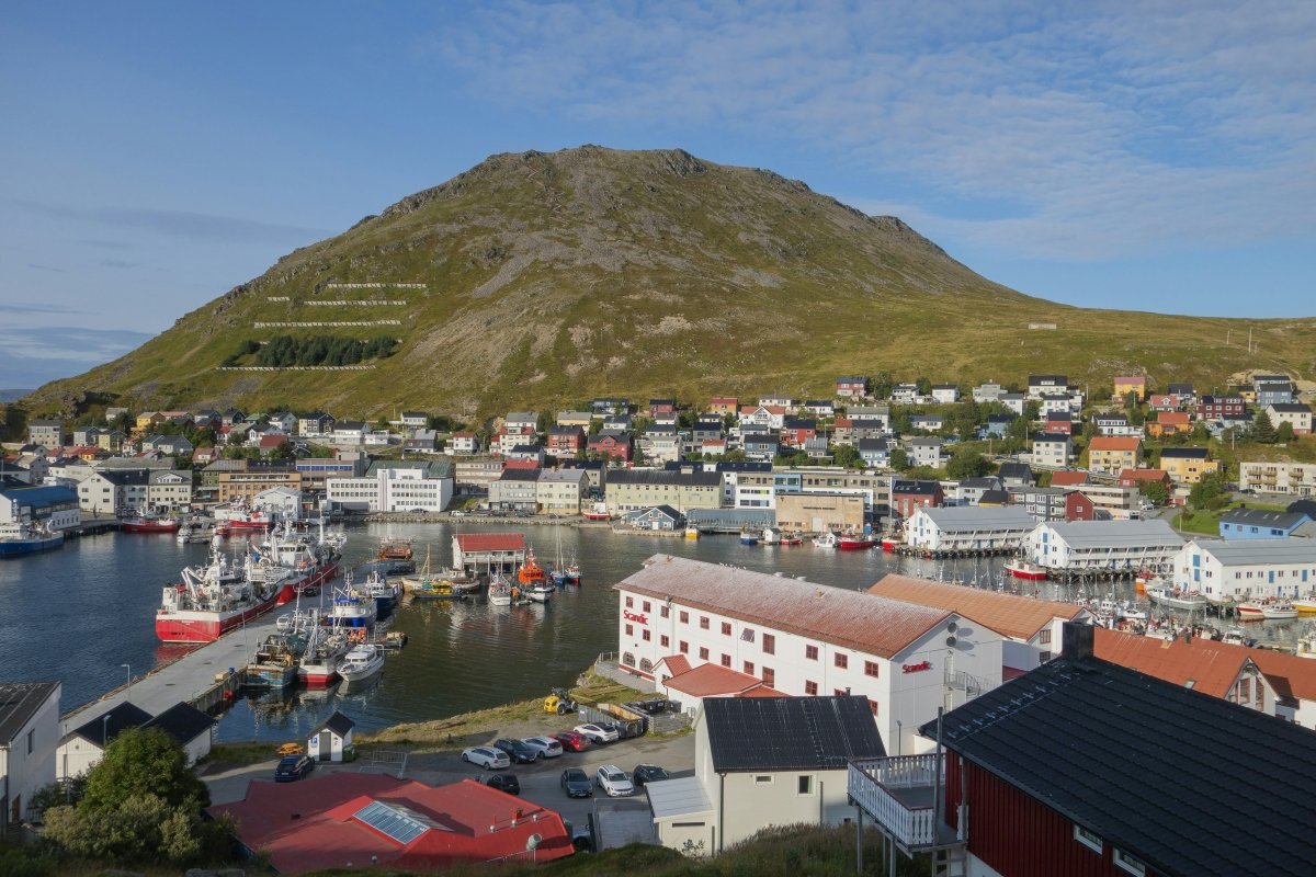 A picturesque view of a coastal town with colorful buildings, boats in the harbor, and a mountain in the background under a clear blue sky.