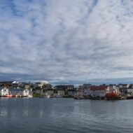 A panoramic view of a coastal harbor featuring fishing boats and colorful buildings against a backdrop of blue sky and clouds.