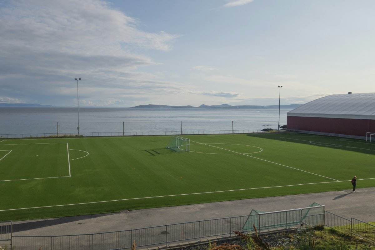 A panoramic view of a well-maintained soccer field adjacent to a tranquil sea, with distant islands visible under a partly cloudy sky.