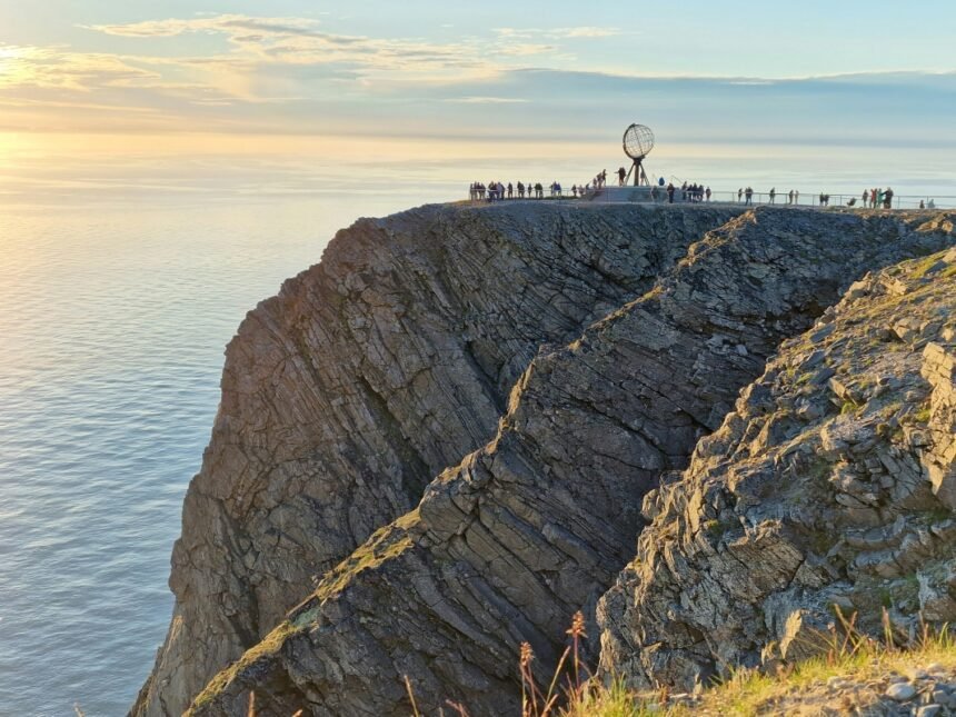 A breathtaking view of the Nordkapp cliff, with visitors gathered near the globe monument overlooking the serene ocean during sunset.