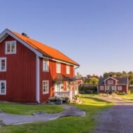A picturesque view of a red wooden house with a vibrant roof, surrounded by greenery and other charming houses under a clear blue sky.