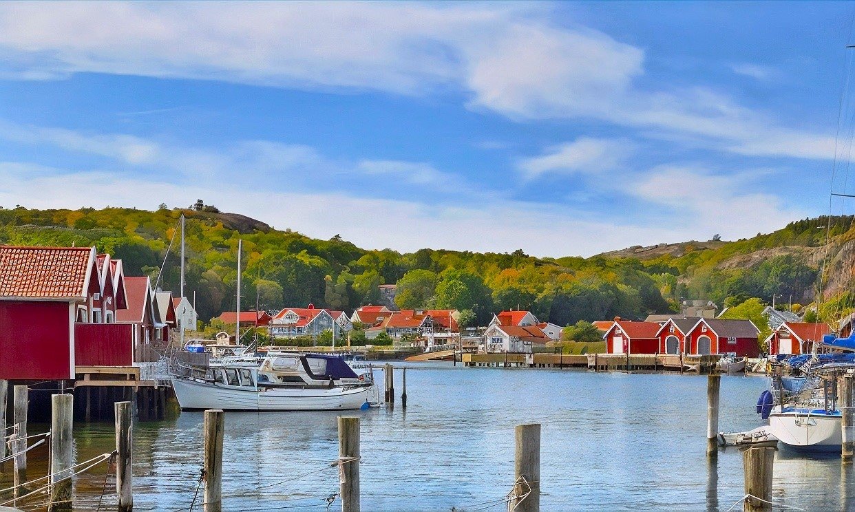 A serene view of a coastal town featuring red-roofed houses, boats docked in the water, and lush green hills under a clear blue sky.