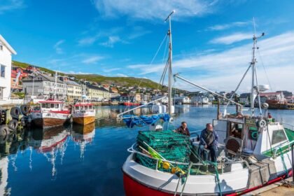 Two fishermen relax on a boat in a tranquil harbor surrounded by colorful buildings and serene waters.